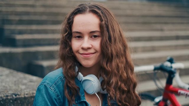 Close up of the teen Caucasian girl with long hait and big headphones on her neck smiling to the camera with her bicycle on the steps outside. Portrait.