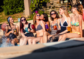 Group of girlfriends at a poolside summer party sitting at the edge of a swimming pool drinking and having fun.