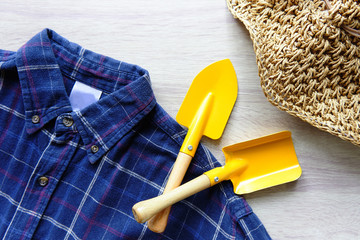 Gardening equipment include a yellow shovel trowel, plaid shirt and hat, wood background.