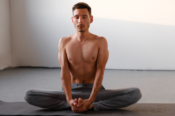 Athletic young man with a naked torso meditating alone. The guy practices yoga in the lotus position indoors, the concept of freedom and tranquility, close-up