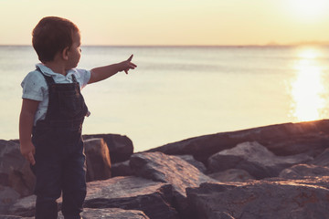 Toddler pointing up with his hand while posing in seaside during sunset with warm colors