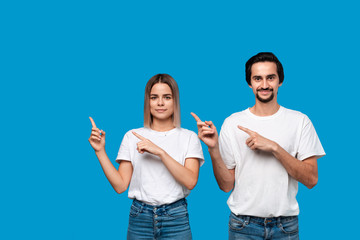 Happy couple in white t-shirts and jeans pointing at the left corner with fingers standing isolated over blue background.
