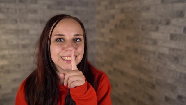 A young woman in a red hoodie is doing silencing gesture with index finger at her lips on a gray brick background. The concept of silence.