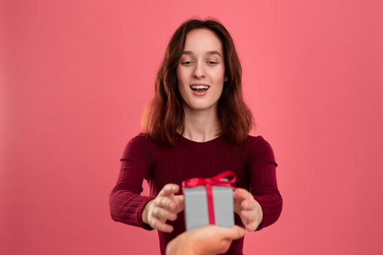 Happy pretty brunette girl gets a present box with a ribbon from another person standing isolated on a dark pink background and smiling at the camera. Celebrating special event.