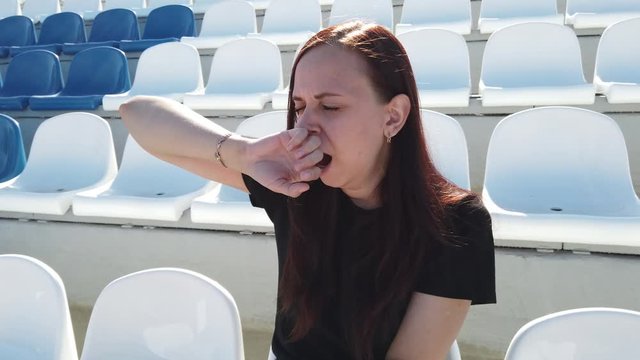 A young woman in black clothes with long hair sits on a stadium bleachers alone and yawns from fatigue on a sunny day.