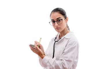 Caucasian young female doctor serius face with glasses and stethoscope prescribing in a notebook on white background isolated