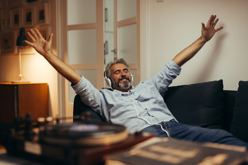 mid aged man listening music with headphones on record player, relaxed in sofa at his home