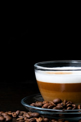 Transparent glass cup hot cappuccino with visible layers of coffee, milk and foam and coffee beans on black wood table at cafe. Dark scene. Place for text. Stylish toning, smoke