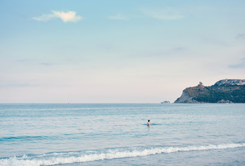 Fototapeta premium a boy into the water looking yacht and calm seascape view of ( sella del diavolo ) in the City of Cagliari from Poetto beach - Sardinia - Italy.