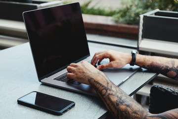 Young tattooed man working on laptop in a coffee shop. Rear view of hipster hands with tattoo busy...