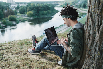 Young millennial student with dreadlocks sitting on grass and using portable laptop computer...