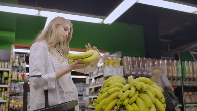 Young beautiful brunette girl in her 20's picking out bananas and putting them into shopping cart at the fruit and vegetable aisle in a grocery stor.