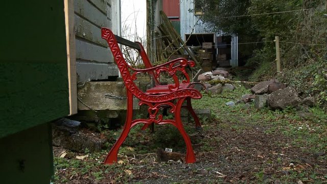 Wide shot showing a red metal bench outside an old farmhouse with tin barns in the background