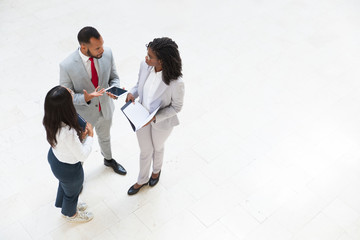 Diverse business colleagues discussing work issues in office hallway. Business man and women standing in circle, holding tablet and papers and talking. Corporate meeting concept