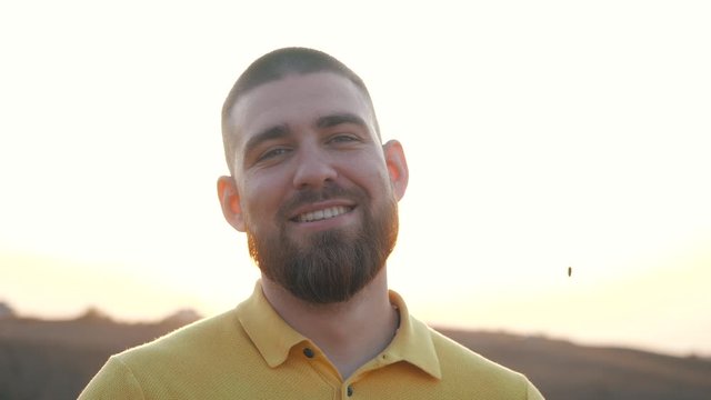 Close up, Portrait of a handsome Caucasian bearded young man looking to the camera and smiling on the background of a bright sunset over natural landscape. Joy of life, active lifestyle, blogger.