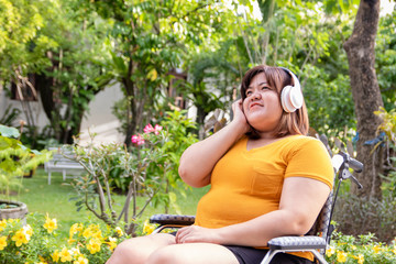 Chubby woman listening to music with headphones on a wheelchair. She has high blood pressure, resulting in paralysis and her has bright smile in the garden. Therapy concept
