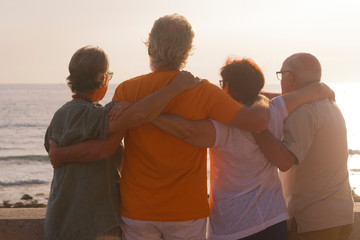 group of seniors hugged together at the beach looking at the sea with a beautiful sunset - family...