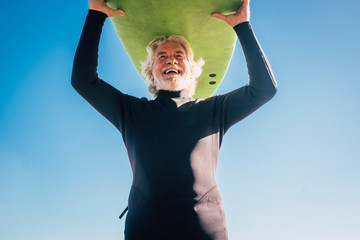 happy senior with surftable on his head is smiling and laughing - old and mature man having fun surfing with a black wetsuits - active retired adult doing activity alone - Powered by Adobe