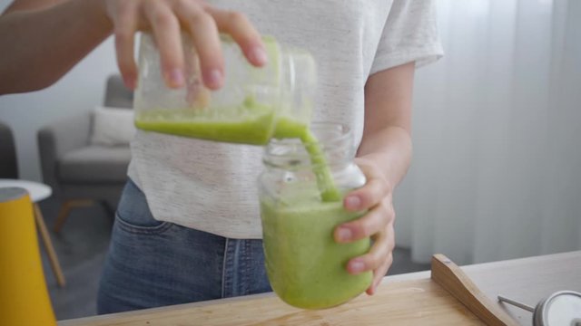 Young slim woman pouring smoothie from the shaker in the small jar in the kitchen at home. Concept of healthy food. Profession of nutri therapist, nutritionist, wellness coach. Camera moving closer