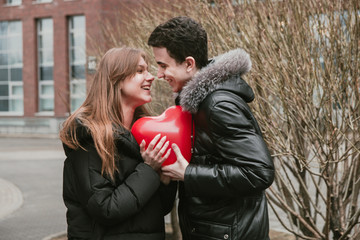 Fototapeta na wymiar A cute couple of young people in love holding a red ball in the shape of a heart, hugging each other and experiencing tender feelings, Valentine's Day