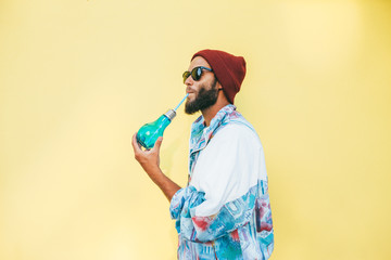 Stylish young hipster man with beard in red hat and a retro jacket of 90s on yellow background. Hipster guy drinks a cocktail.