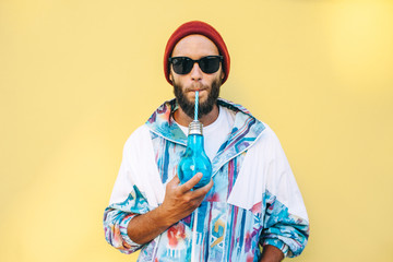 Stylish young hipster man with beard in red hat and a retro jacket of 90s on yellow background. Hipster guy drinks a cocktail.