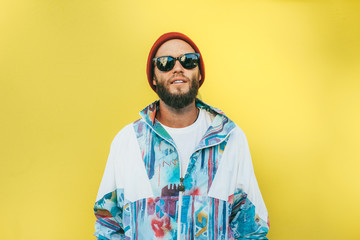 Stylish young hipster man with beard in red hat and a retro jacket of 90s on yellow...