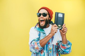 Stylish young hipster man with beard in red hat, sunglasses and a retro jacket of 90s with VHS cassette on yellow background.