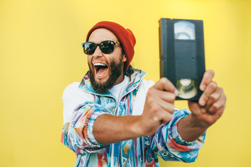 Stylish young hipster man with beard in red hat, sunglasses and a retro jacket of 90s with VHS...