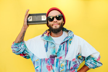 Stylish young hipster man with beard in red hat, sunglasses and a retro jacket of 90s with VHS...