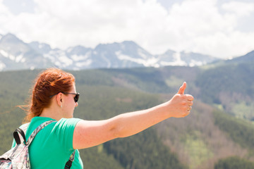 Young woman with backpack giving thumbs up. Trekking and tourism concept. Copyspace. A young pretty redheaded woman standing on a background of mountains