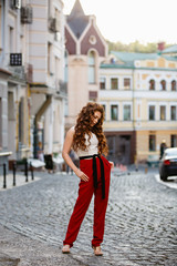 Young curled girl weared in classic trouser jumpsuit on background of old City. Fashion style girl with long curled red hair. Autumn or early spring in the City. Street style Modern strict urban girl.