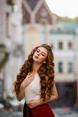 Fototapeta na wymiar Young curled girl weared in classic trouser jumpsuit on background of old City. Fashion style girl with long curled red hair. Autumn or early spring in the City. Street style Modern strict urban girl.
