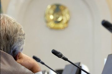 An official with gray hair supports his head with his hand. Blurred background for Kazakhstan news. Microphones, monitors and emblem. The concept of state problems or the solution of popular issues.