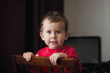 Cute 2 year old boy in a red shirt posing standing on a chair.