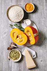 Obraz na płótnie Canvas Ingredients for cooking pumpkin risotto. Raw uncooked risotto rice in ceramic bowl, sliced pumpkin, cream, seeds, parmesan cheese, garlic and herbs over grey wooden background. Flat lay, space
