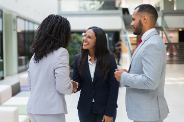 Female diverse business partners shaking hands with each other in hallway. Business man and women standing in office hall, talking and smiling. Successful partnership concept