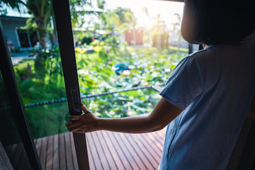 A woman who wearing a blue dress wakes up in the morning, opens the curtain and the glass door. The sunrise at the resort has a natural, beautiful lotus pond on a relaxing day from her tiring work.