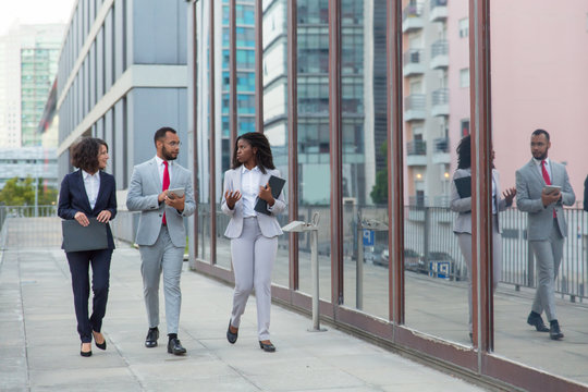 Professional Multiethnic Business Team On Street. Serious Business Colleagues With Folders And Digital Tablet Walking Together Outside Office Building. Cooperation Concept