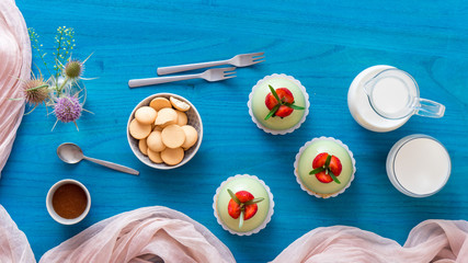Blue wooden table with 3 no-bake strawberry mini sphere matcha cheesecakes with bowl of salt caramel and buiscuits, glass pitches and glass of milk.