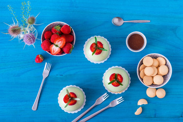 Blue wooden table with 3 no-bake strawberry mini sphere matcha cheesecakes with bowl of salt caramel and buiscuits, glass pitches and glass of milk.