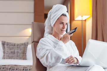 Young girl student talking on smartphone with her family relaxing in hotel room after shower in towel and bathrobe looking laptop and social network. Millennials vacation concept in foreign countries