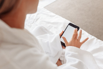 Unidentified young woman in a white bathrobe looks at a smartphone while sitting on a bed on an early weekend morning. Time management concept. Business management. View social networks and mail
