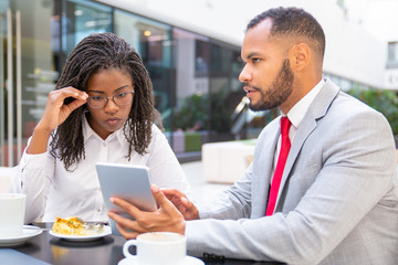Serious business man showing project presentation to colleague. Business man and woman sitting in cafe, using tablet together and talking. Teamwork concept
