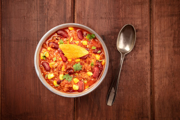 Chili con carne, a Mexican stew with red beans, cilantro leaves, ground beef, and chili peppers, overhead shot with a nacho chips on a dark rustic wooden background