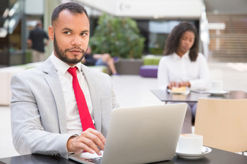 Positive businessman working on laptop in co-working space. Young handsome man in office suit sitting at table in cafe, using computer and looking away. Wi-Fi concept