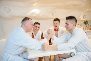 close up photo of 4 men in white gowns sitting around a table and drinking beer after sauna procedures
