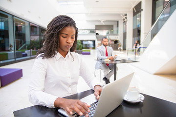 Serious female professional working on project during coffee break n office hall. Young African American business woman using laptop, man drinking coffee in background. Wireless technology concept