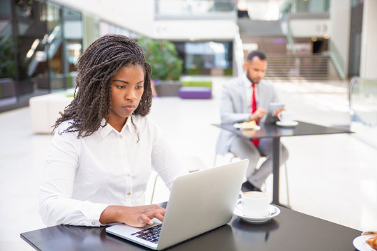Focused hardworking female professional working on report during coffee break. Young African American business woman using laptop, man using tablet in background. Co-working concept