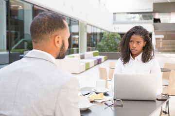 Young African American office employee having lunch with colleague. Business man and woman sitting at table in cafe, using laptop and talking. Corporate communication concept